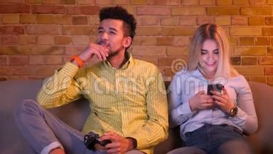 非洲男人<strong>专心</strong>地用操纵杆玩电子游戏，他漂亮的白种人女朋友在舒适的环境中看电话